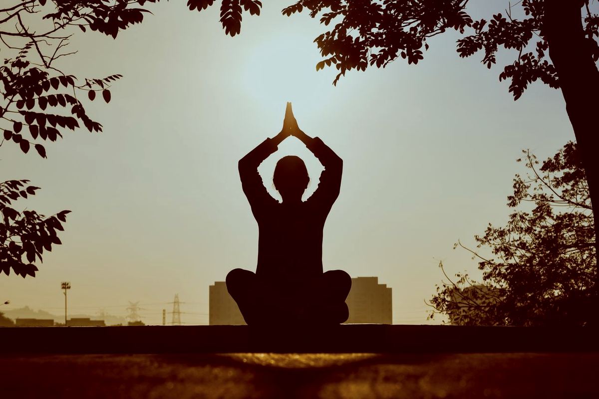 A woman seated outdoors in a calm yoga pose at sunset, hands raised in a restorative stretch